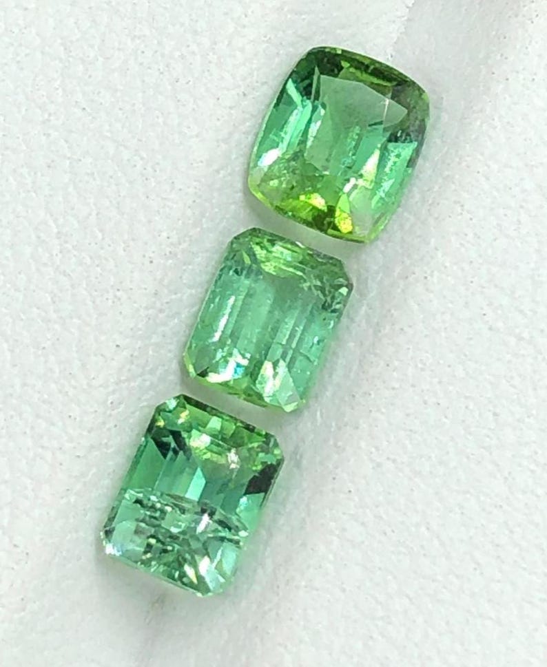 May include: Three faceted, green gemstones with a rectangular shape. The gems have a vibrant, light green colour with a slight yellow hue. The gemstones are displayed on a white surface.