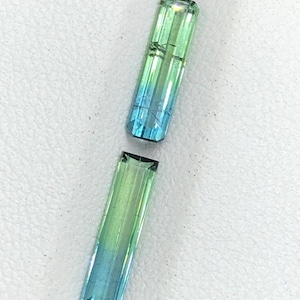 May include: Two rectangular tourmaline gemstones. The gemstones transition from green to blue. The facets of the stones are clearly visible, reflecting light. The stones are set against a white background.