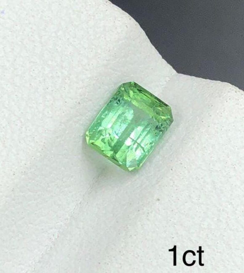 May include: A rectangular, light green gemstone with visible internal structures. The gem is set against a white background and the text "1ct" is visible in the lower right corner. The gemstone appears to be faceted.