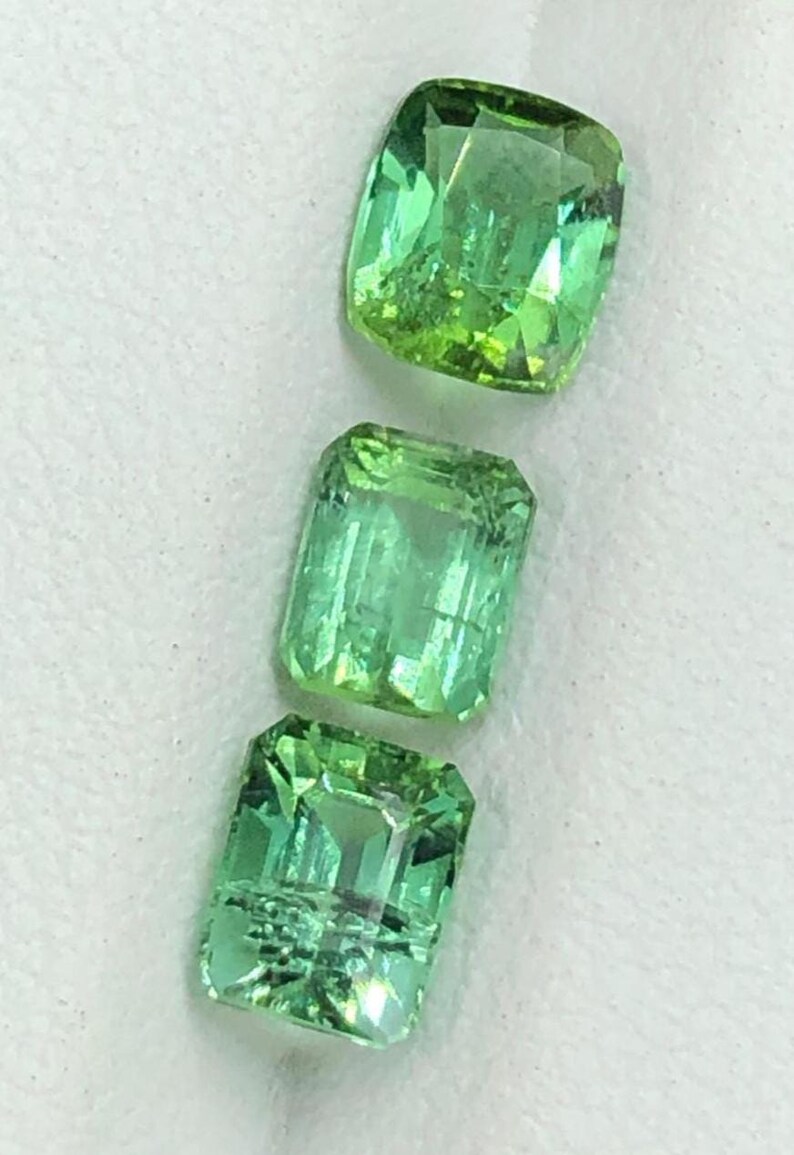 May include: Three faceted, green gemstones are displayed against a white background. The gems have a rectangular and cushion cut. The gems are a vibrant, light green colour, and reflect light.