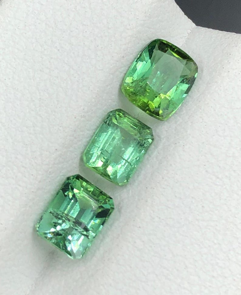 May include: Three bright green gemstones, cut in rectangular and cushion shapes, are set against a white backdrop. The gems are translucent, reflecting light and revealing their internal structure. The stones vary in shade, from lighter to deeper green.
