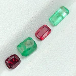 May include: Four gemstones: two emeralds and two rubies. The emeralds are a vibrant green, while the rubies are a deep red. Each gem is rectangular with faceted surfaces, reflecting light. The gems are arranged diagonally on a white surface.