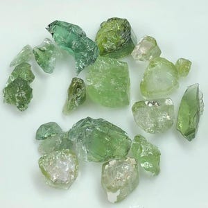 Puede incluir: Una colección de piedras preciosas en bruto, sin tallar, en tonos verdes y transparentes. Las piedras varían en tamaño y forma, con una textura rugosa y natural. La paleta de colores va desde verdes pálidos y translúcidos hasta tonos más profundos y opacos.