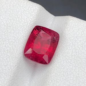 Puede incluir: Una piedra preciosa de rubí tallada en cojín. La gema es de un color rojo intenso y saturado con inclusiones internas visibles. La piedra se encuentra sobre un fondo blanco texturizado, resaltando su brillo y color.