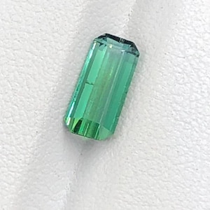 May include: A rectangular, emerald-green gemstone with a faceted cut. The gem has a translucent quality, with visible internal striations. The stone is set against a white background, highlighting its vibrant color and clarity. The gem is likely a tourmaline.