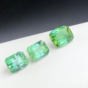 May include: Three vivid green gemstones with a rectangular cut are shown against a white and black backdrop. The gems have a slight sparkle, suggesting they are polished. The stones are likely for jewellery making or collecting.