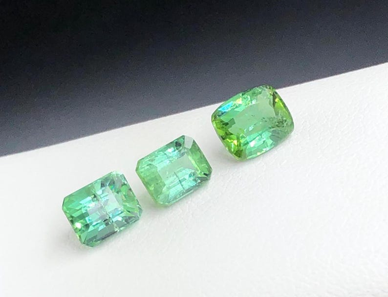 May include: Three vivid green gemstones with a rectangular cut are shown against a white and black backdrop. The gems have a slight sparkle, suggesting they are polished. The stones are likely for jewellery making or collecting.