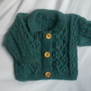 May include: A teal green knitted cardigan with a shawl collar and three wooden buttons. The cable knit design features textured patterns. The baby sweater is a cozy, handmade garment.