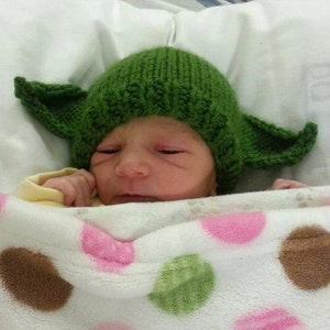 Könnte beinhalten: Ein neugeborenes Baby trägt eine grüne gestrickte Yoda-Mütze und liegt auf einer weißen Decke mit rosa und braunen Punkten.
