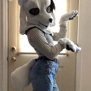 May include: A white dog fursuit head with large, dark eyes and floppy ears. The fursuit is worn with a striped long-sleeve shirt and ripped blue jeans. A white tail is attached to the jeans. The fursuit is a fun costume.
