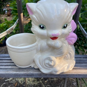 May include: A vintage ceramic planter shaped like a white kitten with pink accents. The kitten has green eyes, a pink nose, and a red mouth. A light pink bow is on the back. The planter has a small, round opening on the side.