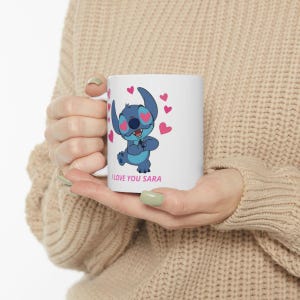 K&ouml;nnte beinhalten: Wei&szlig;e Keramik-Tasse mit einer blauen Cartoon-Figur mit herzf&ouml;rmigen Augen und rosa Herzen. Auf der Tasse steht der Text "I LOVE YOU SARA". Die Tasse wird von einer Person gehalten, die einen beigen Pullover tr&auml;gt.