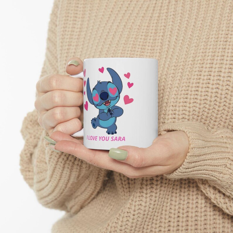 K&ouml;nnte beinhalten: Wei&szlig;e Keramik-Tasse mit einer blauen Cartoon-Figur mit herzf&ouml;rmigen Augen und rosa Herzen. Auf der Tasse steht der Text "I LOVE YOU SARA". Die Tasse wird von einer Person gehalten, die einen beigen Pullover tr&auml;gt.