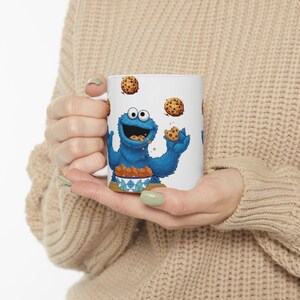 Puede incluir: Taza de cerámica blanca con el Monstruo de las Galletas de Barrio Sésamo. El personaje azul está rodeado de galletas con chispas de chocolate, algunas en un cuenco y otras flotando. La taza la sostiene una persona.