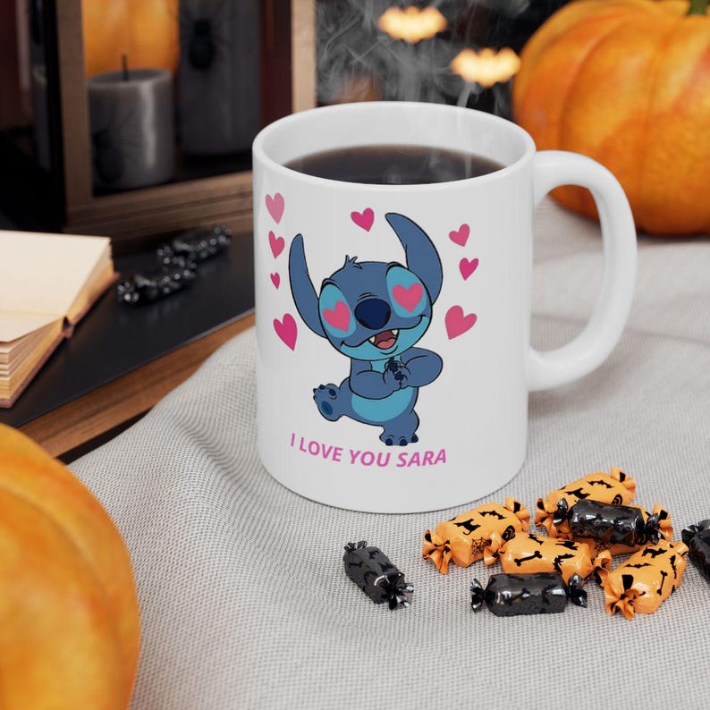 K&ouml;nnte beinhalten: Wei&szlig;e Keramik-Tasse mit einer blauen Cartoon-Figur und rosa Herzen, mit dem Text "I LOVE YOU SARA". Die Tasse enth&auml;lt eine dunkle Fl&uuml;ssigkeit. Halloween-Dekorationen im Hintergrund.