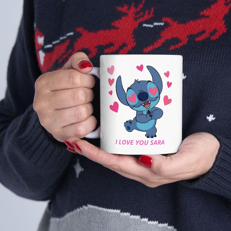 K&ouml;nnte beinhalten: Wei&szlig;e Keramik-Tasse mit einer blauen Cartoon-Figur, rosa Herzen und dem Text "I LOVE YOU SARA". Die Tasse hat einen Henkel und wird von einer Person gehalten, die einen dunkelblauen Pullover tr&auml;gt.