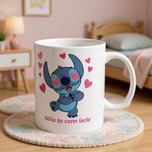 K&ouml;nnte beinhalten: Wei&szlig;e Keramik-Tasse mit einer blauen Cartoon-Figur, herzf&ouml;rmigen Akzenten und rosa Herzen. Auf der Tasse steht der Text "Ni&ntilde;a to cann lecle". Die Tasse steht auf einer cremefarbenen Matte mit floralem Design.
