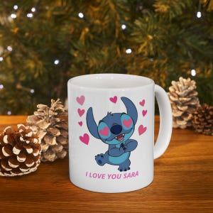 K&ouml;nnte beinhalten: Wei&szlig;e Keramiktasse mit einem blauen Cartoon-Charakter, herzf&ouml;rmigen Augen und rosa Herzen. Der Text "I LOVE YOU SARA" ist rosa gedruckt. Die Tasse hat einen wei&szlig;en Henkel und steht auf einer Holzoberfl&auml;che.