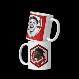 May include: Two white ceramic mugs with red and white designs. The top mug features a black and white illustration of a soccer player with a red and white soccer team logo. The bottom mug features a red and white soccer team logo with a black and white illustration of a soccer player.