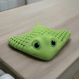 May include: A light green, cartoon-style stingray figurine with large, black-pupiled eyes. The stingray has a textured, scale-like pattern across its body and a small, pointed tail. The figurine is resting on a light-colored wooden surface.