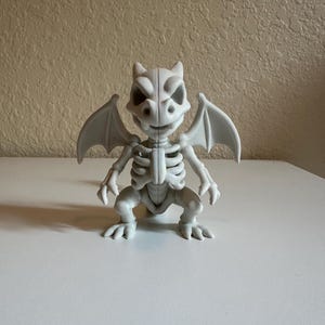 Peut inclure: Figurine grise claire d'un squelette de gargouille avec des ailes, debout sur une surface blanche. La figurine pr&eacute;sente des caract&eacute;ristiques squelettiques d&eacute;taill&eacute;es, notamment des c&ocirc;tes, une colonne vert&eacute;brale et un cr&acirc;ne avec de grandes orbites. Les ailes sont d&eacute;ploy&eacute;es.