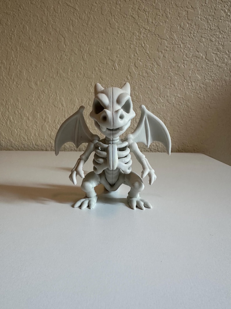 Peut inclure: Figurine grise claire d'un squelette de gargouille avec des ailes, debout sur une surface blanche. La figurine pr&eacute;sente des caract&eacute;ristiques squelettiques d&eacute;taill&eacute;es, notamment des c&ocirc;tes, une colonne vert&eacute;brale et un cr&acirc;ne avec de grandes orbites. Les ailes sont d&eacute;ploy&eacute;es.
