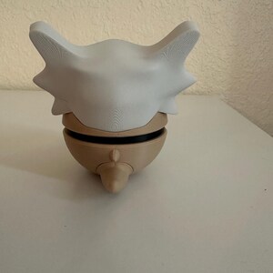 May include: A 3D-printed Cubone skull mask in white and tan. The mask features a large, curved bone-like structure with pointed edges and a rounded base. The mask is designed to fit over the head.