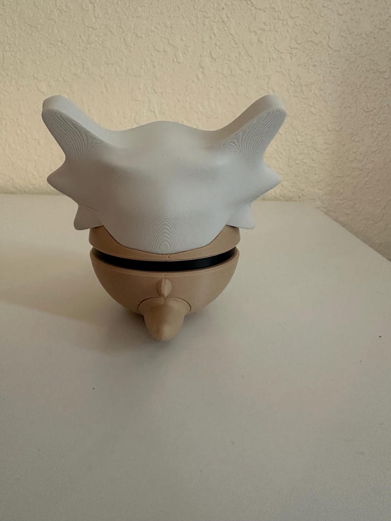 May include: A 3D-printed Cubone skull mask in white and tan. The mask features a large, curved bone-like structure with pointed edges and a rounded base. The mask is designed to fit over the head.