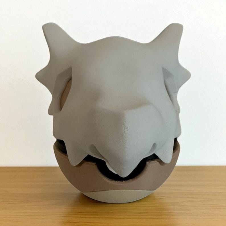 May include: A gray, 3D-printed Cubone skull resting on a brown and black base. The skull has pointed ears and a detailed texture. The base is a rounded shape, and the overall design is a stylized representation of a character.