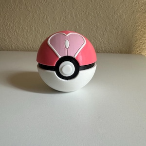 Puede incluir: Un ratón de ordenador rosa, blanco y negro con forma de Poké Ball. El ratón tiene un diseño en forma de corazón en la parte superior y un botón blanco. El ratón está sobre una superficie blanca.