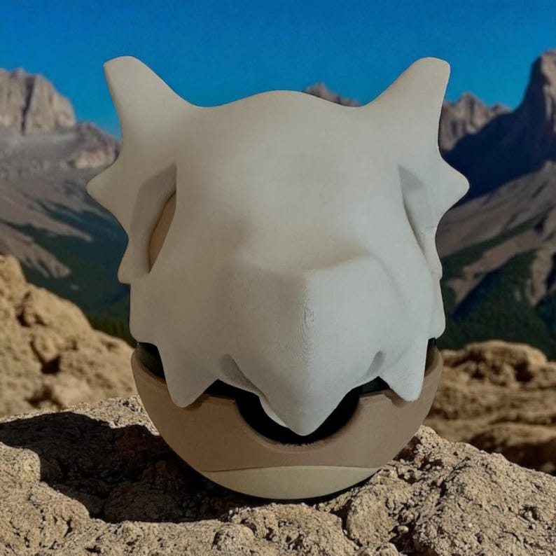 May include: A white, stylized skull-shaped object with pointed ears and a rounded base. The base is tan and brown, with a black opening. The object is set against a backdrop of mountains and a blue sky.