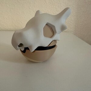 May include: A white Cubone skull mask resting on a tan and black spherical base. The mask has a smooth, matte finish with detailed bone structure, including eye sockets and a prominent brow ridge. The base appears to be a stylized Pok&eacute; Ball.