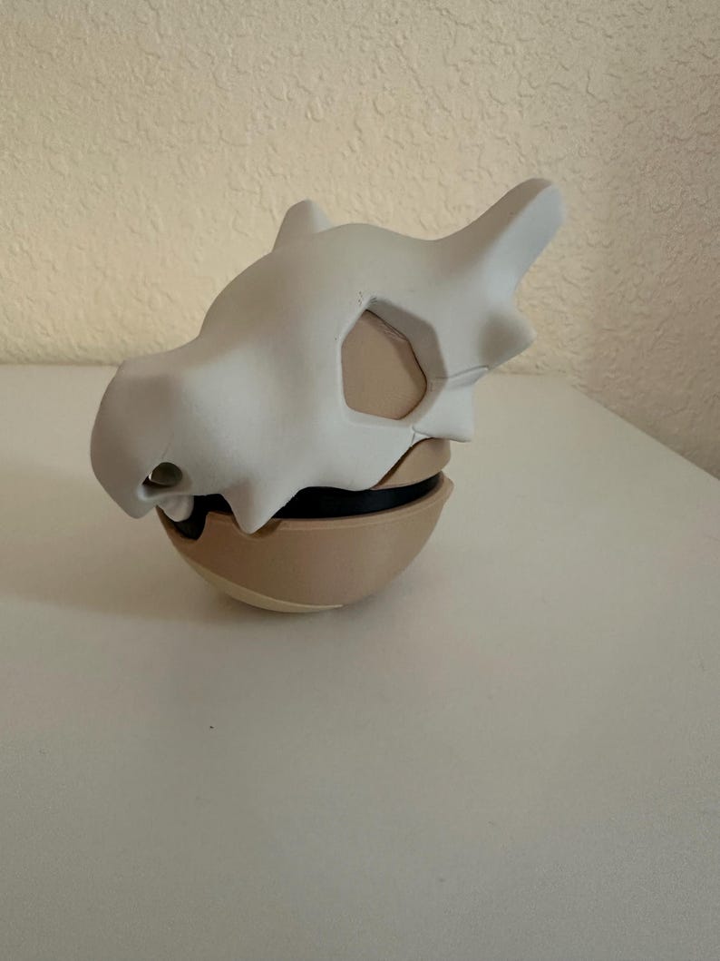 May include: A white Cubone skull mask resting on a tan and black spherical base. The mask has a smooth, matte finish with detailed bone structure, including eye sockets and a prominent brow ridge. The base appears to be a stylized Pok&eacute; Ball.
