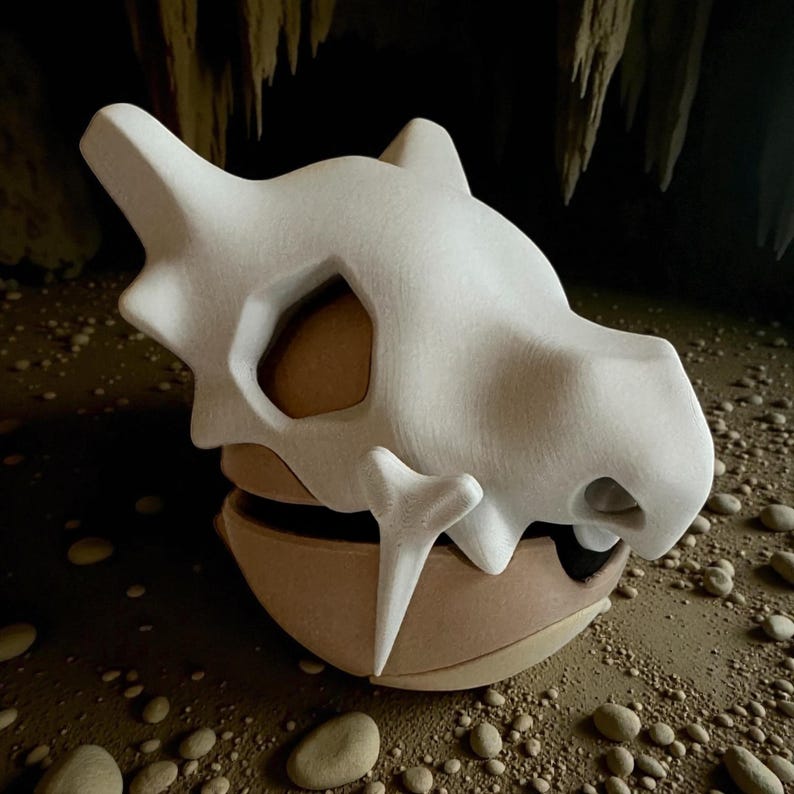 May include: A white, stylized skull-shaped sculpture with a brown base. The sculpture has large eye sockets and a prominent jawline. The background features a cave-like setting with rocks and stalactites.