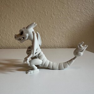 Peut inclure: Figurine de dragon blanc imprim&eacute;e en 3D avec un design squelettique. Le dragon est pos&eacute; avec ses pattes avant pli&eacute;es, sa queue enroul&eacute;e et ses ailes d&eacute;ploy&eacute;es. La figurine est en mat&eacute;riau clair et repose sur une surface blanche.