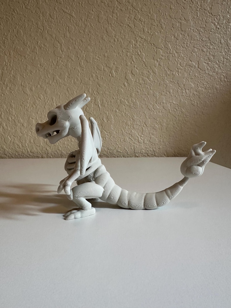 Peut inclure: Figurine de dragon blanc imprim&eacute;e en 3D avec un design squelettique. Le dragon est pos&eacute; avec ses pattes avant pli&eacute;es, sa queue enroul&eacute;e et ses ailes d&eacute;ploy&eacute;es. La figurine est en mat&eacute;riau clair et repose sur une surface blanche.