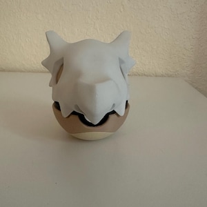 May include: A Cubone skull mask, white in color, sits atop a tan and brown egg-shaped object. The mask has pointed ears and a prominent brow ridge. The object is on a white surface.