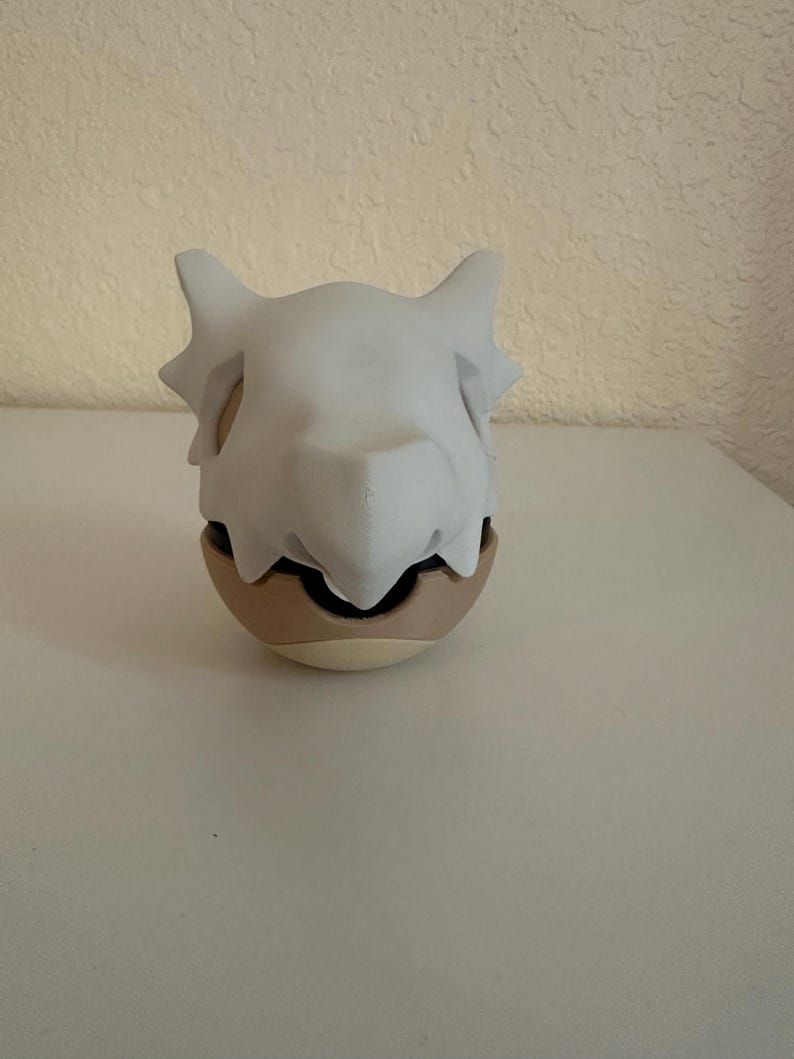 May include: A Cubone skull mask, white in color, sits atop a tan and brown egg-shaped object. The mask has pointed ears and a prominent brow ridge. The object is on a white surface.