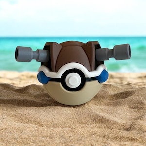 May include: A brown and gray Poké Ball toy with blue accents. The Poké Ball is on a sandy beach.