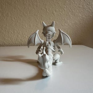 Peut inclure: Figurine blanche imprim&eacute;e en 3D repr&eacute;sentant une cr&eacute;ature ressemblant &agrave; un dragon, avec des ailes de chauve-souris, des cornes et une longue queue. La figurine est en position accroupie, les ailes d&eacute;ploy&eacute;es. Les d&eacute;tails de la structure squelettique de la cr&eacute;ature sont visibles.
