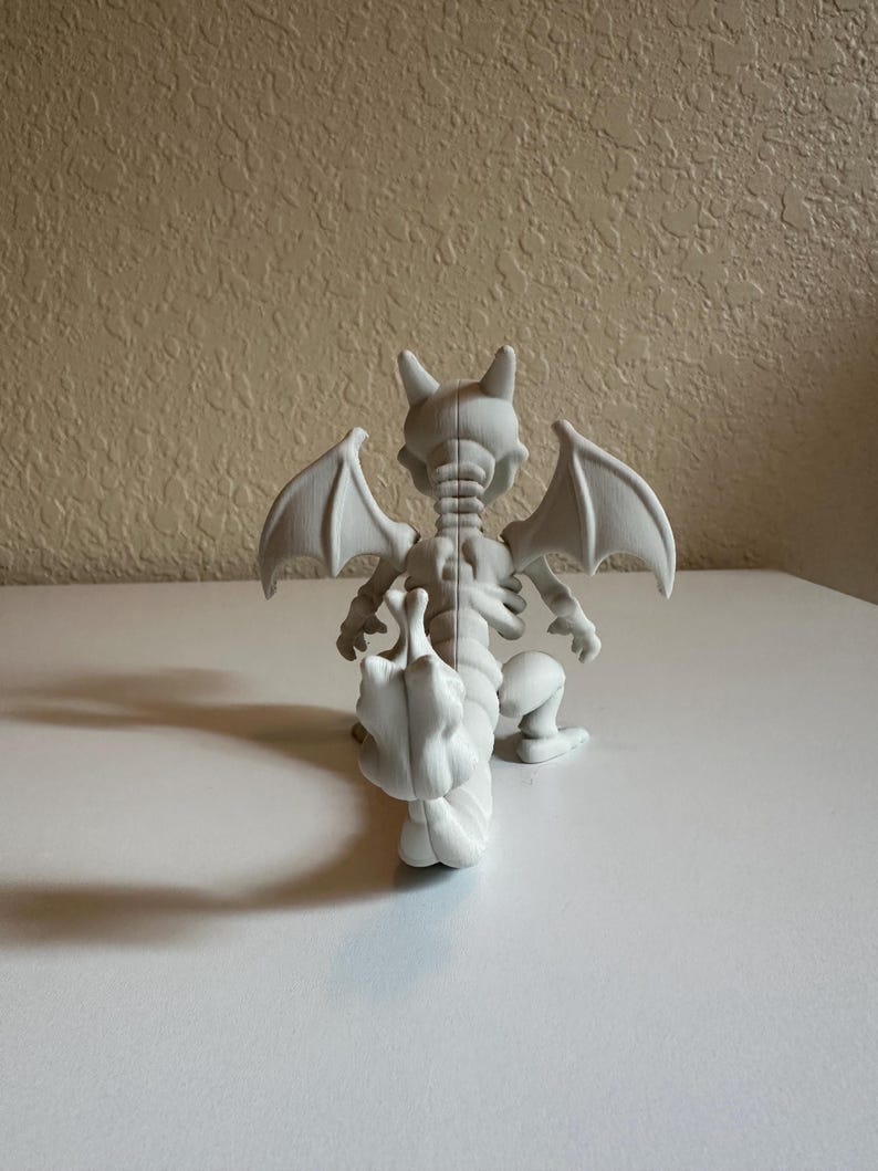 Peut inclure: Figurine blanche imprim&eacute;e en 3D repr&eacute;sentant une cr&eacute;ature ressemblant &agrave; un dragon, avec des ailes de chauve-souris, des cornes et une longue queue. La figurine est en position accroupie, les ailes d&eacute;ploy&eacute;es. Les d&eacute;tails de la structure squelettique de la cr&eacute;ature sont visibles.