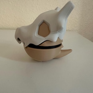 May include: A Cubone skull mask, featuring a white bone-like exterior and a tan interior. The mask has a rounded base and a black band. The design is inspired by the popular Pok&eacute;mon franchise.