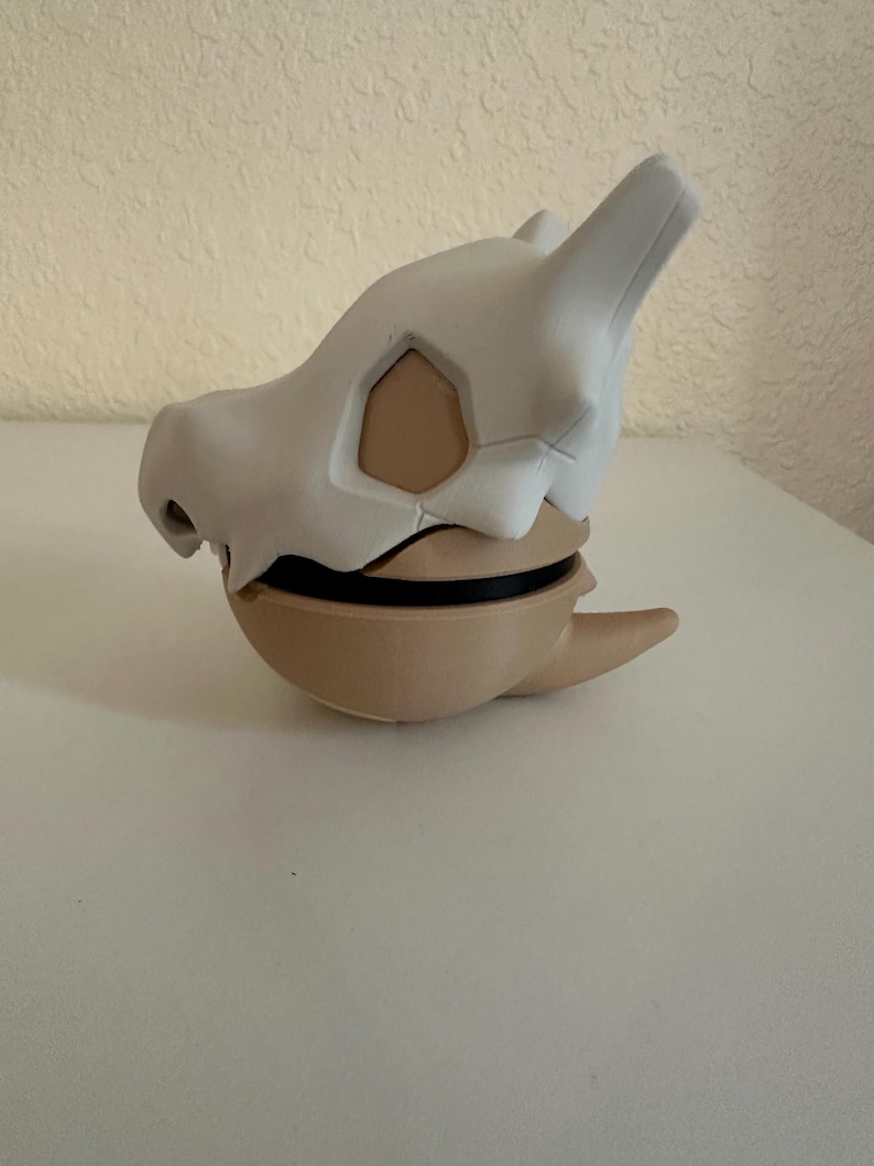 May include: A Cubone skull mask, featuring a white bone-like exterior and a tan interior. The mask has a rounded base and a black band. The design is inspired by the popular Pok&eacute;mon franchise.