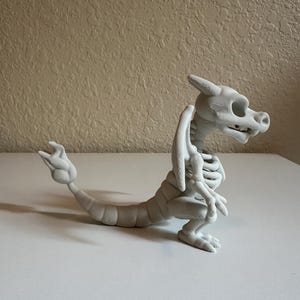 Peut inclure: Figurine de dragon squelettique gris clair imprim&eacute;e en 3D. Le dragon est en position assise, la queue tendue. La figurine pr&eacute;sente des d&eacute;tails sur la cage thoracique, les ailes et la t&ecirc;te. Sur une surface blanche.