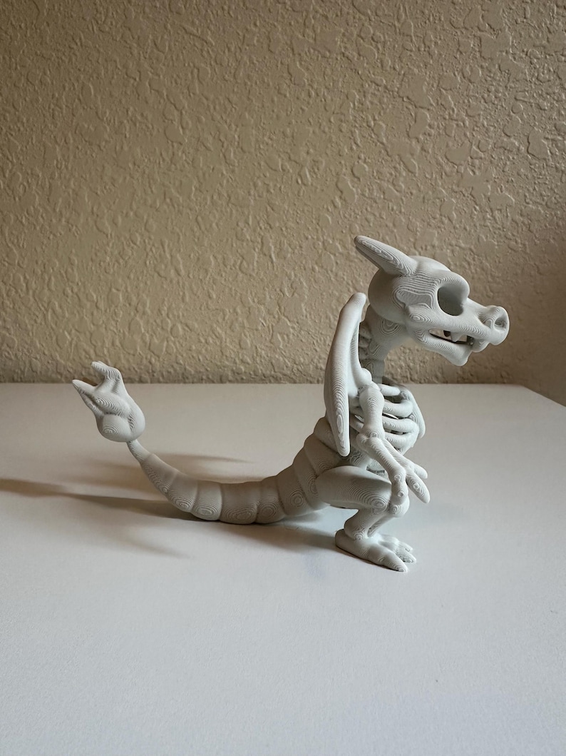 Peut inclure: Figurine de dragon squelettique gris clair imprim&eacute;e en 3D. Le dragon est en position assise, la queue tendue. La figurine pr&eacute;sente des d&eacute;tails sur la cage thoracique, les ailes et la t&ecirc;te. Sur une surface blanche.