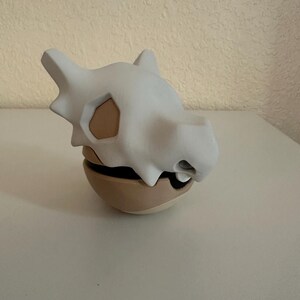 May include: A white Cubone skull-shaped container with a tan base. The skull has eye sockets and a snout. The container is a two-piece design, with the skull resting on the base. The overall design is a stylized representation of the Pok&eacute;mon Cubone.