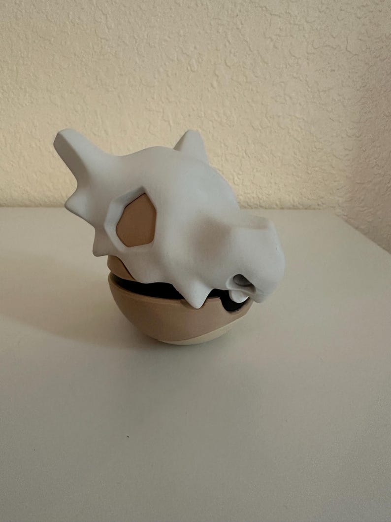 May include: A white Cubone skull-shaped container with a tan base. The skull has eye sockets and a snout. The container is a two-piece design, with the skull resting on the base. The overall design is a stylized representation of the Pok&eacute;mon Cubone.