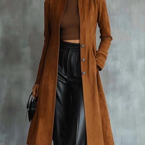 Peut inclure: Un long manteau marron à texture suédée doté d'un col montant, de manches longues et d'une fermeture à boutons sur le devant. Le manteau est porté sur un haut marron à col montant et un pantalon noir. Un petit sac à main noir est tenu sur le côté.