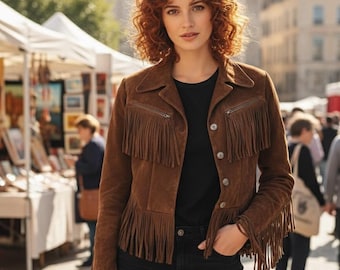 Puede incluir: Chaqueta de ante marrón con flecos en el pecho, las mangas y la espalda. La chaqueta tiene una parte delantera abotonada y un cuello. La persona de la imagen lleva una camisa negra y vaqueros negros. Una bolsa de cuero marrón está en la mano de la persona.