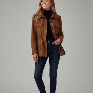 Pode incluir: Casaco de camurça castanho com quatro bolsos frontais e cintura com cordão. O casaco é usado com uma gola alta preta e jeans azul escuro. A pessoa está usando botas marrons.